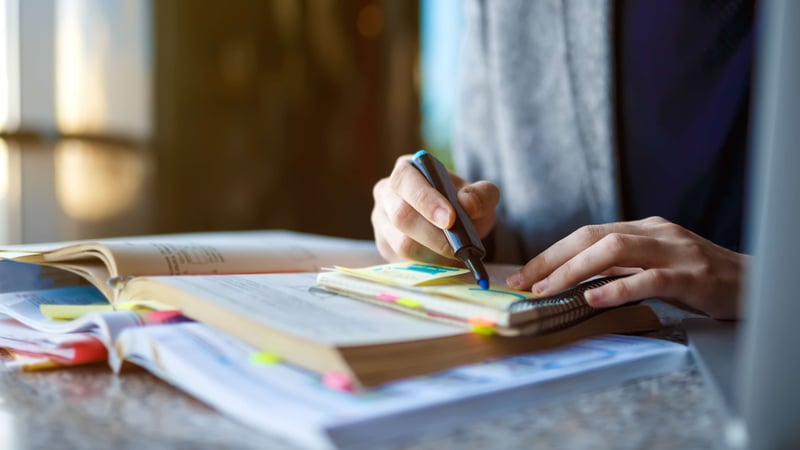 Close-up of a hand marking up a sticky note with a highlighter, surrounded by colour-coded tabs on an open book, showing the trial and error of working through a problem by hand rather than reaching for a finished answe