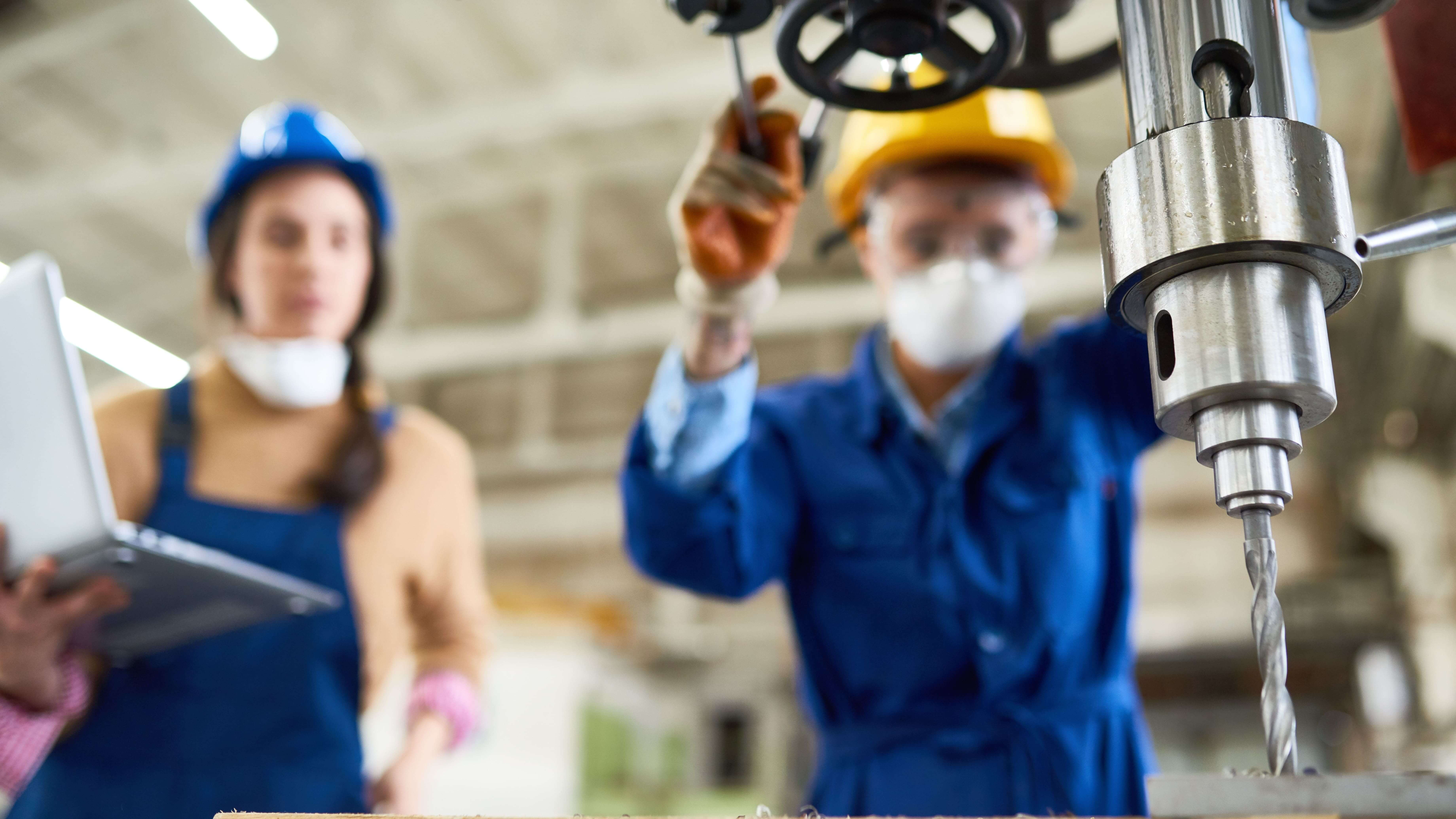 A supervisor in a blue hard hat observes a worker operating a large industrial drill press