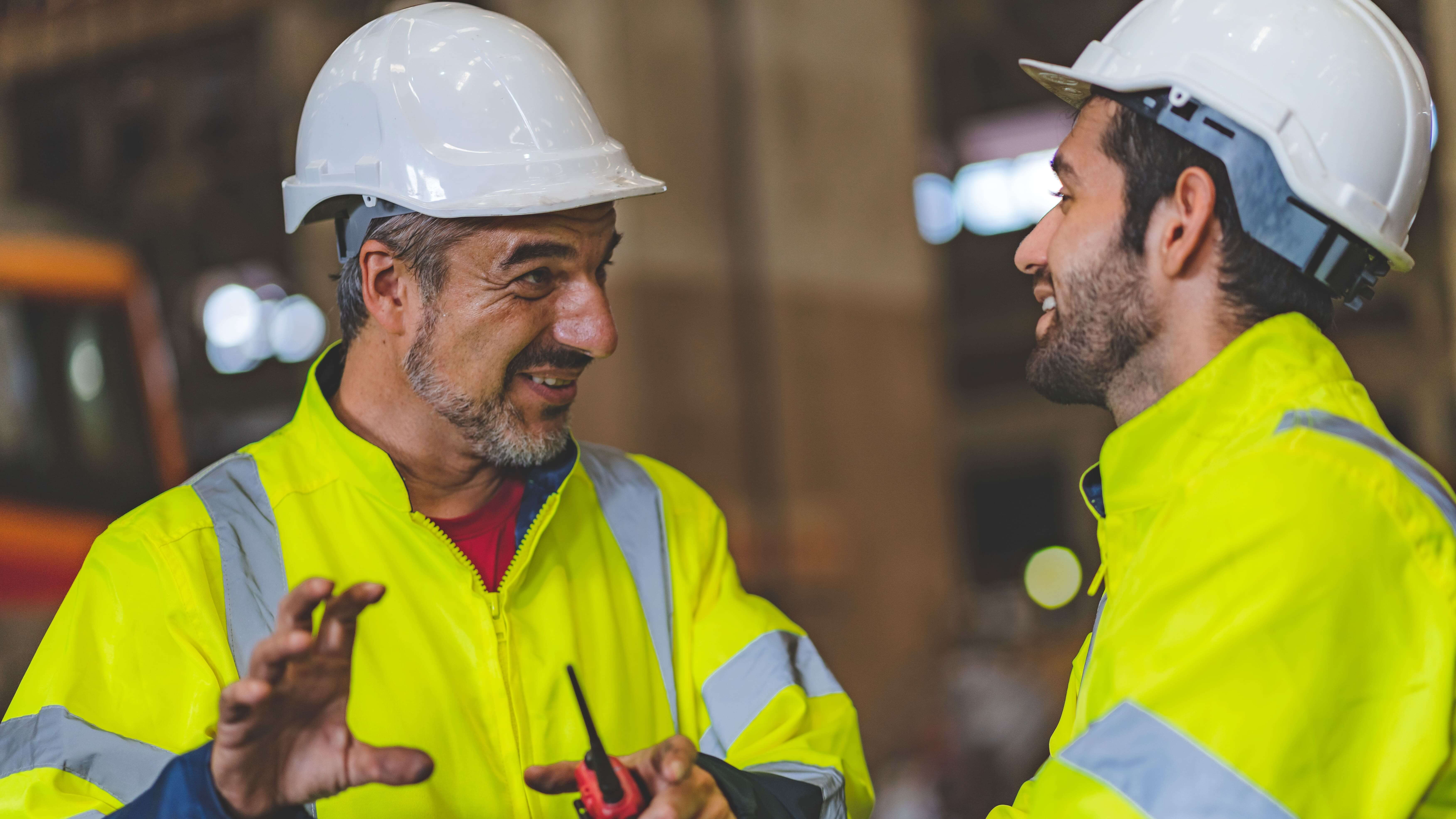 An experienced industrial worker wearing a hard hat and high-visibility jacket provides coaching to a younger colleague during a shift